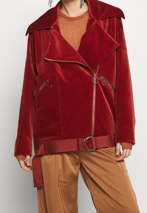 Kobieta w czerwonej aksamitnej kurtce z suwakami, dużym kołnierzem i paskiem, w połączeniu z rdzawej barwy swetrem i jasnobrązowymi spodniami.