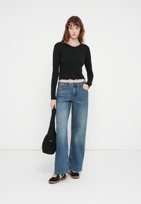 Cardigan scurt cu mânecă lungă, negru, cu nasturi, asortat cu blugi evazați, de culoare albastru deschis. Modelul ține o geantă moale, neagră și poartă adidași negri.