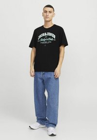 Sort bomulds T-shirt med hvid og turkis tekst, der viser "JACK & JONES Surfer's Club 1990." Sammen med blå lige ben jeans og hvide sneakers.