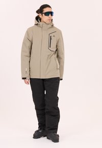 Beige vattentät jacka med hög krage och svarta accenter, matchad med svarta byxor och svarta skidstövlar. Innehåller en dragkedjeförsedd bröstficka.