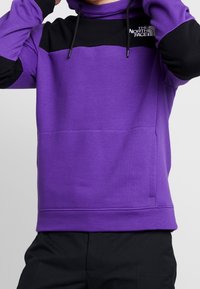 Lila sweatshirt med svarta accenter, som har huva, framficka och broderad logotyp. Slät textur med ribbade muddar. Justerbara snoddar.