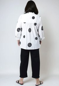 Witte blouse met zwarte bloemborduursels, losvallend model, lange mouwen, geplisseerde achterkant, gecombineerd met zwarte geplooide broek en sandalen.