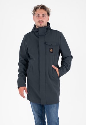 Giacca impermeabile grigia scura con colletto alto, tasche frontali e un patch con logo sul lato sinistro del petto, con una finitura liscia e texture.