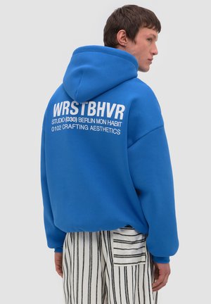 Junger Mann im Profil, trägt einen blauen Kapuzenpullover mit weißem Schriftzug und gestreifte weiß-schwarze Hose vor schlichtem Hintergrund.