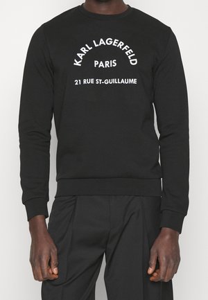 Person trägt ein schwarzes Langarmshirt mit weißem Text „Karl Lagerfeld Paris 21 Rue St-Guillaume“ auf der Vorderseite.