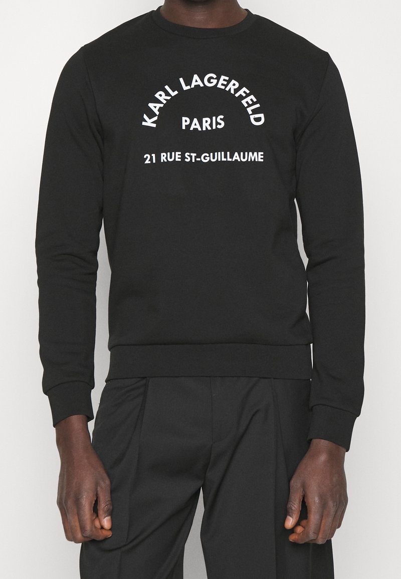 Personne portant un t-shirt noir à manches longues avec un texte blanc indiquant "Karl Lagerfeld Paris 21 Rue St-Guillaume" sur le devant.
