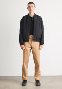 Black bomber jacket with textured fabric, zip front, and ribbed cuffs, worn over a black polo and tan trousers, paired with black shoes.