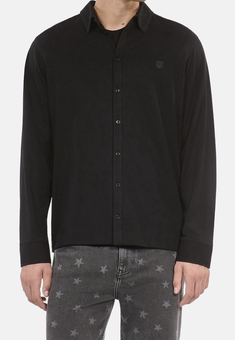 Homme portant une chemise noire à manches longues boutonnée associée à un jean gris avec un motif étoile délavé, debout devant un fond uni.