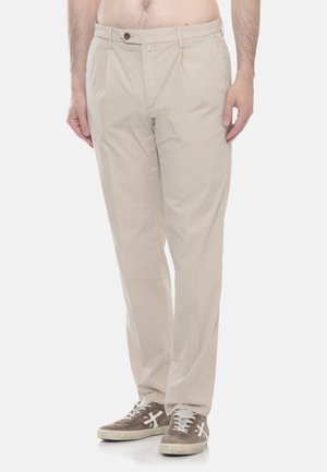 Uomo con pantaloni beige e sneaker marroni, in piedi davanti a uno sfondo bianco, mani rilassate lungo i fianchi.