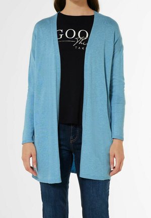 Femme portant un cardigan bleu clair ouvert sur un t-shirt noir graphique et un jean bleu foncé, debout devant un fond blanc.