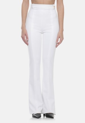 Donna che indossa pantaloni bianchi a vita alta a zampa d'elefante, su misura, con cintura coordinata e décolleté nere con punta affilata, in piedi davanti a uno sfondo semplice.