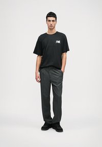 T-shirt preto de algodão com um gráfico branco no lado esquerdo do peito, combinado com calças cinza-escuro riscas e sapatos pretos. O modelo está de pé.