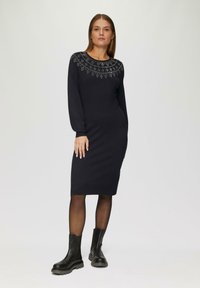 Schwarzes Strickkleid mit langen Ärmeln und Rundhalsausschnitt, mit aufwendigen silbernen Verzierungen am Ausschnitt. Ein Paar schwarze Knöchelstiefel.