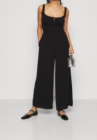 Kvinna klädd i en svart ärmlös jumpsuit med breda ben, svarta Mary Jane-skor och håller en liten silverhandväska i höger hand.