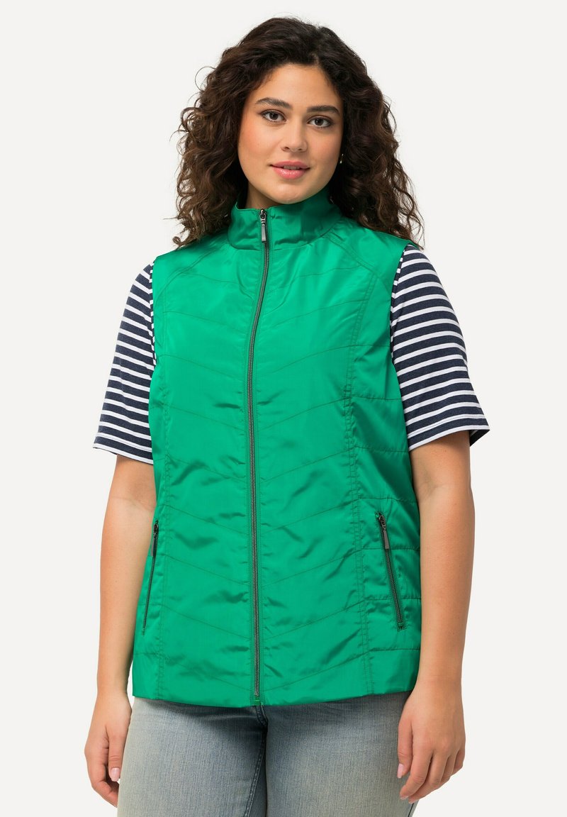 Groen gewatteerd vest met een hoge kraag, ritssluiting en twee zijzakken. Gedragen over een marineblauw en wit gestreept T-shirt met korte mouwen.