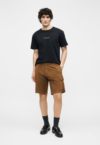 Jeune homme aux cheveux bouclés portant un T-shirt noir, un short cargo marron, des chaussettes foncées et des mocassins en cuir noir, debout devant un fond blanc.