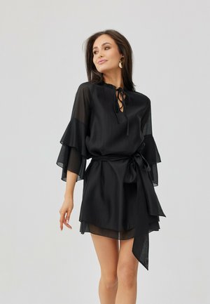 Žena v černých průsvitných dlouhých rukávech s volánky na rukávech a zavazováním v pase, nosící zlaté náušnice, stojící před světle šedým pozadím.