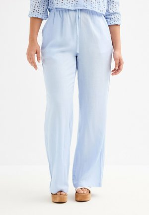 Mujer con pantalones de pierna ancha azul claro con cintura ajustable con cordón y sandalias de plataforma, de pie sobre un fondo blanco.