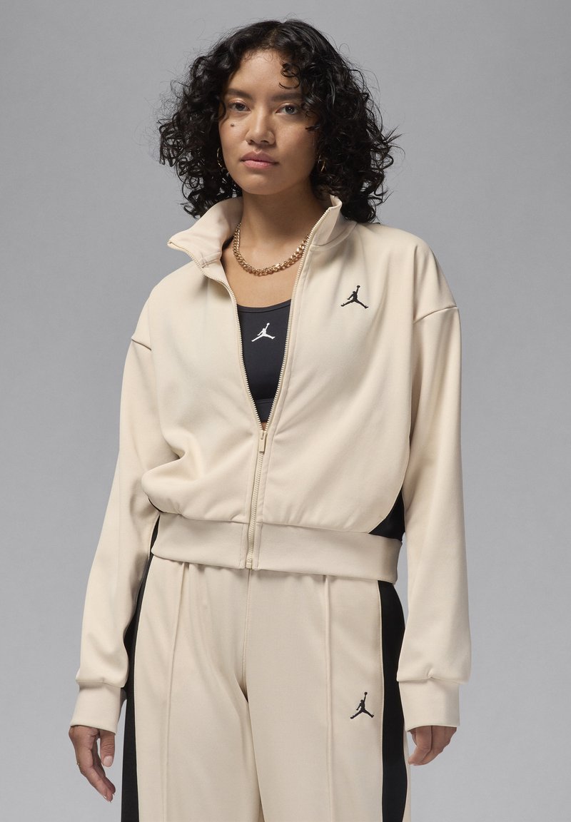 Veste de survêtement beige zippée et pantalon assorti avec des panneaux latéraux noirs, ornés de logos noirs brodés. Tissu lisse et athlétique, coupe décontractée.