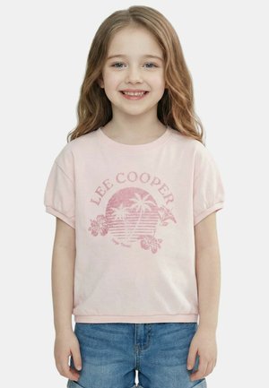 Fille souriante aux longs cheveux portant un T-shirt Lee Cooper rose clair avec un motif de palmier et un short en jean, debout devant un fond blanc.