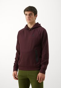 Sweat-shirt à capuche bordeaux avec une poche zippée à l'avant, des cordons réglables et un logo noir sur la poitrine ; associé à un pantalon vert.