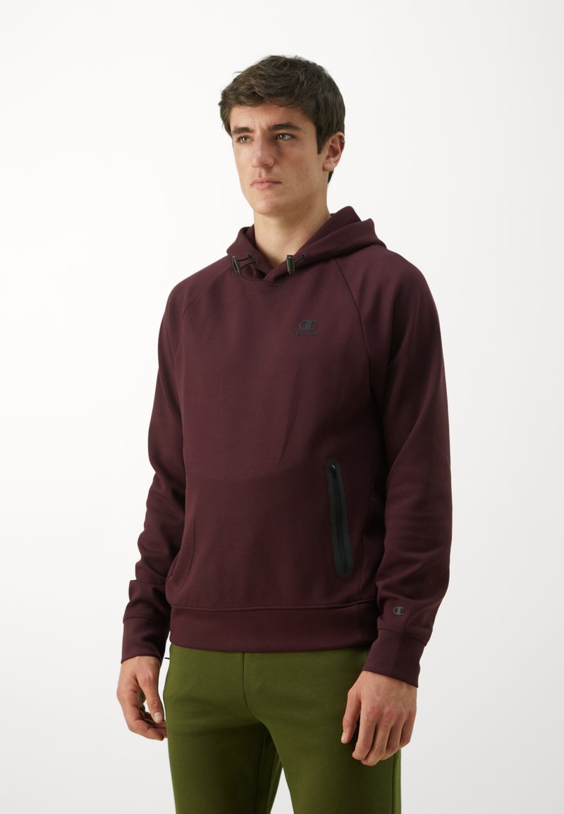 Sweat-shirt à capuche bordeaux avec une poche zippée à l'avant, des cordons réglables et un logo noir sur la poitrine ; associé à un pantalon vert.