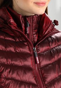 Mørkerød puffjakke med høj krave, lynlås foran og med logo på lynlåsene. Har en glat, blank overflade og quiltet design.