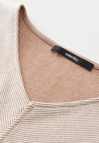 Zbliżenie dzianiny prążkowanej z wąskimi białymi i beżowymi paskami oraz czarną metką odzieżową z napisem „someday” na beżowym wnętrzu kołnierza.