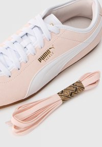 Ροζ σουέντ αθλητικό παπούτσι με λευκές λεπτομέρειες, διαθέτει υφή επιφάνειας, επίπεδα λευκά κ ορδόνια και χρυσό λογότυπο. Περιλαμβάνει επίσης ροζ κορδόνια που ταιριάζουν.