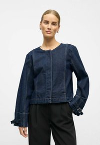 Dunkelblaue Denimjacke, cropped Design, runder Ausschnitt, lange Ärmel mit verstellbaren Riemen, sichtbare Nähte, getragen über schwarzen Hosen.