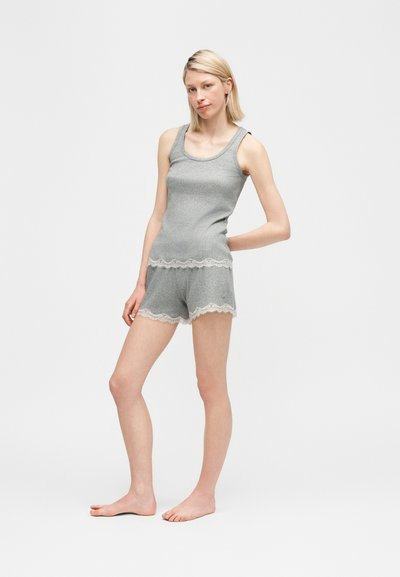 Νεαρή γυναίκα στέκεται ξυπόλυτη φορώντας γκρι ριγωτό τοπ χωρίς μανίκια και ταιριαστό σορτς με λευκή δαντέλα σε απλό φόντο.