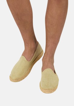 Osoba nosząca beżowe wsuwane espadryle z plecioną podeszwą z juty, stojąca na tle jednolitego, jasnego tła.