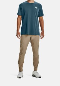 Mans blåa tränings t-shirt med logotyp på bröstet, tillsammans med beige byxor med fickor och en smal passform. Vita sneakers fulländar outfiten.