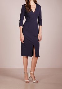 Mörkblå figursydd klänning med V-ringad hals, trekvartslånga ärmar och sprund framtill. Kombinerad med vita klackskor med ankelsnodd och scallop-detaljer.
