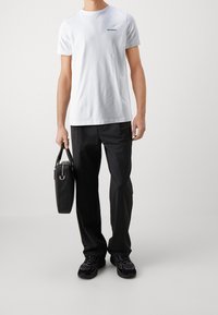 Vit bomull t-shirt med tryckt logo, parat med svarta byxor och svarta sneakers. Håller en svart läderväska med metalldetaljer.