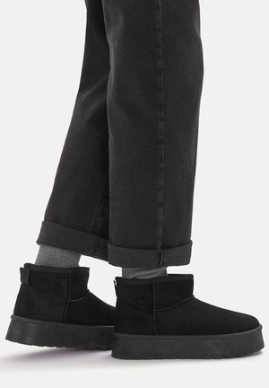 Botines de ante negro con punta redonda, con suela de goma plana y una lengüeta trasera para facilitar ponerlos. Llevados con pantalones negros remangados.