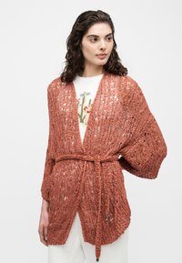 Γυναίκα που φοράει πλεκτή ζακέτα σε σκουριά απόχρωση με ζώνη πάνω από ένα λευκό τοπ με λουλουδάτο σχέδιο, στέκεται μπροστά σε ένα απλό, ανοιχτόχρωμο φόντο.