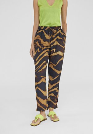 Pantalones a rayas de tigre en negro y naranja con cinturilla elástica, bolsillos laterales y detalles de cordón, combinados con chanclas verdes brillantes.