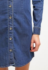 Denim mekko, jossa on napitus edessä, pitkät hihat ja kaareva helma. Kankaassa on keskimäinen sininen sävy ja sileä rakenne.
