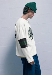 Biela dlhá košeľa s zelenými a čiernymi pruhmi, s grafickým potlačou a voľným strihom, spárovaná s modrými džínsami a zelenou čiapkou.