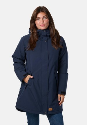 Une femme présente un manteau bleu marine à capuche, avec un col montant et des boutons sur le devant, sur un fond clair et uni.