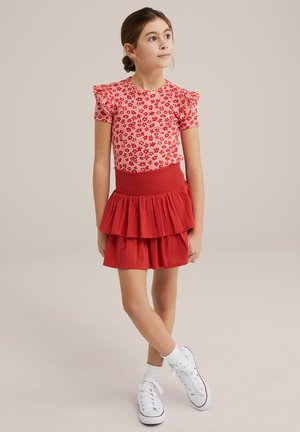 Top corto a maniche corte floreale rosso con spalle arricciate, abbinato a una gonna rossa a strati. Sneakers bianche e calzini alla caviglia completano l'outfit.