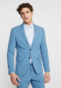 Chaqueta de traje azul claro con dos botones negros, solapas con muesca y bolsillos laterales. Se lleva con una camisa azul claro debajo.