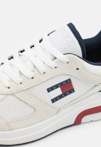 Zapatilla blanca con acentos de ante crema, con suela de goma, cordones redondos y una etiqueta del logo "TOMMY JEANS" en rojo, blanco y azul.