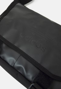 Svart messenger-väska med texturerad, glansig yta. Har en framträdande präglad logotyp, kardborreknäppning och justerbar rem.