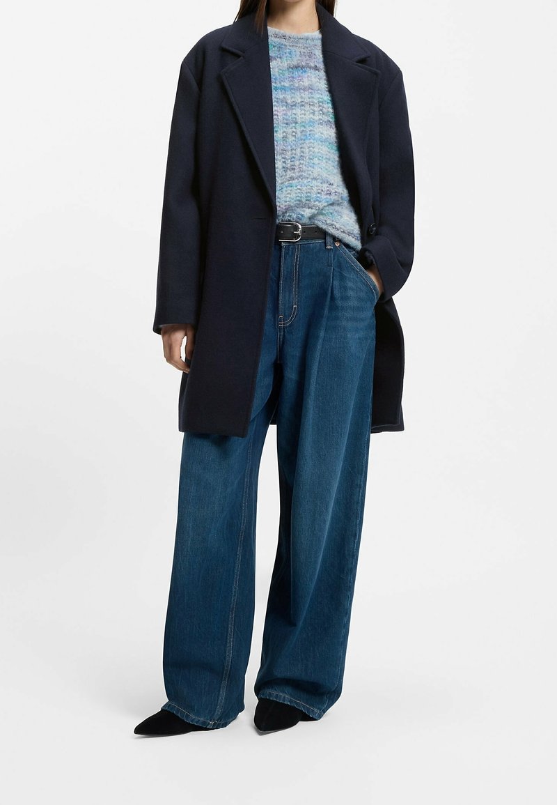 Personne portant un manteau bleu marine foncé par-dessus un pull en maille bleu et gris, un jean bleu à jambes larges et des chaussures noires pointues, debout avec une main dans la poche.