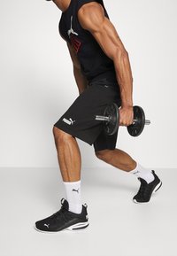 Pantaloni corti sportivi neri, scarpe da ginnastica nere con dettagli bianchi, calze bianche e una maglietta senza maniche nera con logo colorato, mentre tiene un manubrio.