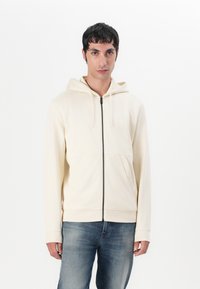 Crème kleurige rits hoodie van zacht materiaal, met een voorvak, een capuchon met trekkoorden en een subtiele logo detail op de borst. G gedragen met blauwe jeans.