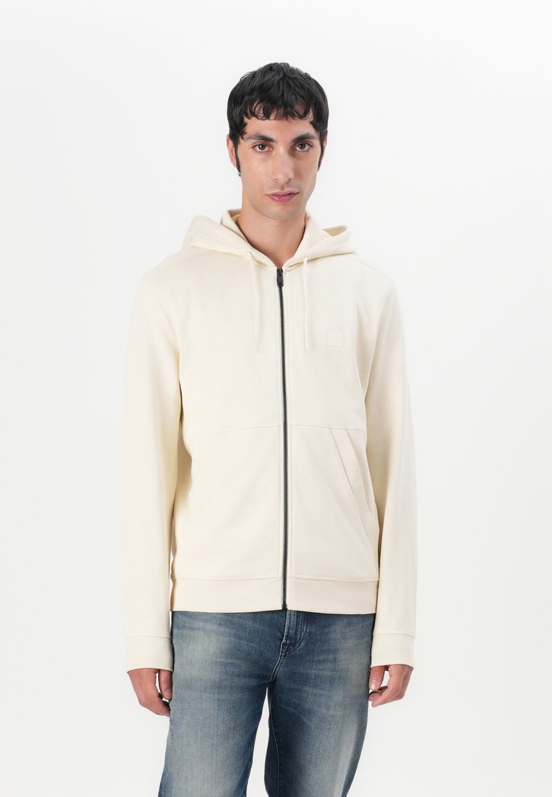 Crème kleurige rits hoodie van zacht materiaal, met een voorvak, een capuchon met trekkoorden en een subtiele logo detail op de borst. G gedragen met blauwe jeans.