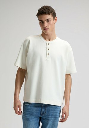 Top blanco de manga corta con cuello redondo y tapeta de tres botones. Tejido texturizado, ajuste relajado, combinado con vaqueros azules.